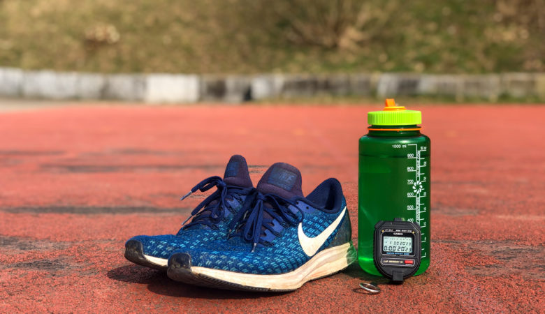 Ein Paar Sportschuhe, eine Trinkflasche und eine Stoppuhr, welche 21 Sekunden anzeigt stehen auf einer Tartanbahn.